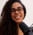 Imagem mostra Lilian, uma mulher negra de pele clara, com cabelos cacheados, que usa óculos de grau e sorri. Ela está com um fone de ouvido e um microfone de gravação de podcast.  Foto: Imagem: Acervo Pessoal/Lilian Moreira / Alma Preta