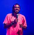 Imagem mostra Jhordan Matheus. Ele é um homem negro, com dreads nos cabelos, sorri e usa uma camisa rosa pink. Em uma das suas mãos, segura um microfone.  Foto: Juliana das Fotos / Alma Preta