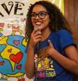 Imagem de divulgação do projeto Favela Mundo. Uma mulher negra e com cabelos cacheados fala ao microfone. Ela veste a camisa do projeto.  Foto: Imagem: Divulgação/ Favela Mundo / Alma Preta