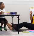 A imagem mostra uma criança negra sentada em uma cadeira e escrevendo em um caderno sobre uma mesa. A criança olha e sorri para uma mulher negra sentada no chão e próxima a uma lousa. Foto ilustra texto sobre livros e filmes para educação antirracista.  Foto: Imagem: Natasha Hall/ Unsplash / Divulgação