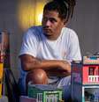 Marcelino é um homem negro que usa dreads. Ele está sentado ao centro da imagem com miniaturas de casas ao redor  Foto: Divulgação/Léu Britto / Alma Preta