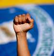 Imagem mostra a bandeira do Brasil ao fundo e a mão fechada de uma pessoa negra para cima.  Foto: Imagem: Reprodução/Sindicato dos Metalúrgicos / Alma Preta