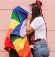 Imagem mostra casal de mulheres se beijando atrás de uma bandeira do arco-íris, símbolo do movimento LGBTQIAP+.  Foto: Imagem: Reprodução/Jéssica Kidermann / Alma Preta