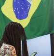 Imagem mostra pessoa utilizando o celular na COP, com a bandeira do Brasil ao fundo.  Foto: Imagem: Pedro Borges/ Alma Preta  / Alma Preta