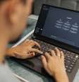 Uma pessoa usa um laptop com o site ChatGPT aberto. Ela é vista parcialmente, de costas e desfocada. As mãos digitam no teclado preto iluminado. Um óculos está sobre a mesa, ao lado do laptop.  Foto: 