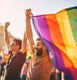 Foto mostra 2 homens em primeiro plano, onde um deles segura a bandeira que representa a luta das pessoas LGBTQIAPN+, durante um evento, com várias outras pessoas desfocadas em segundo plano  Foto: Alma Preta