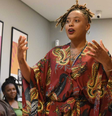 Imagem mostra uma mulher de pele negra, autora do livro ‘Lesbiandade’ centralizada discursando com os braços levemente levantados, em uma sala. A mulher usa um vestido vermelho com detalhes dourados. Ao lado esquerdo, no fundo, mulheres negras sentadas escutando.  Foto: Alma Preta