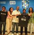 Imagem mostra grupo de pessoas em pé e sorrindo em frente ao banner da campanha "Com racismo não tem jogo", enquanto seguram uma camisa da iniciativa.  Foto: Alma Preta