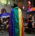 A imagem mostra um homem negro de costa, coberto com a bandeira da população LGBTQIA+  Foto: Alma Preta