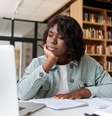 Imagem mostra uma mulher negra, sentada à mesa e olhando para um notebook.  Foto: Alma Preta