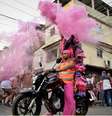 Imagem mostra a performance de Milu Almeida e Luiz Otávio na favela da Nova Holanda (RJ). Na foto, os artistas aparecem em cima de uma moto com trajes na cor rosa e laranja, enquanto dezenas de pessoas os cercam.  Foto: Alma Preta