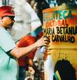 A imagem mostra um homem negro com uma lata de tinta em frente a uma geladeira com os dizeres “Gelateca Cultural Maria Betânia de Carvalho”.  Foto: Alma Preta