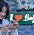 Mulher chinesa checa seu telefone em frente a um letreiro que diz "eu amo Pequim"  Foto: Getty Images / BBC News Brasil
