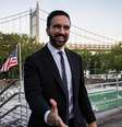 Zohran Mamdani estende a mão. Ele está de terno. Atrás dele, vê-se uma ponte, junto com bandeiras.  Foto: Getty Images / BBC News Brasil