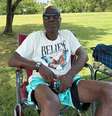 Homem com camiseta branca, shorts azuis e óculos escuros, sentado em uma cadeira de camping sobre a grama  Foto: BBC News Brasil