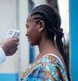Um médico verifica a temperatura de uma mulher usando um termômetro infravermelho sem contato. Somente o braço do médico, que está vestido de branco, é mostrado. A mulher que está com a temperatura verificada tem o cabelo trançado. Ela está vestindo uma blusa azul brilhante com um padrão e está ao lado de uma porta de madeira azul com um prédio branco e azul ao fundo.  Foto: Getty Images / BBC News Brasil