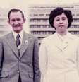 Uma fotografia antiga de Hitomi e seu marido, Charles. Eles estão em pé em frente a uma fonte e um prédio de apartamentos. Hitomi veste um blazer com botões e uma saia. Charles usa um terno cinza com uma gravata listrada. Hitomi está com os braços atrás das costas e Charles com os braços ao lado do corpo. Ambos sorriem e olham diretamente para a câmera. Foto: Hitomi Soga / BBC News Brasil