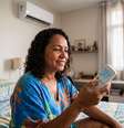 Imagem mostra uma mulher negra de cabelos cacheados sentada na cama com um controle do ar-condicionado na mão. Foto: Imagem gerada por inteligência artificial / Xataka