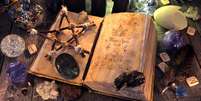 Antigo livro de bruxa com pentagrama, velas pretas, cristais e objetos rituais   Foto: iStock
