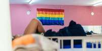 Imagem mostra bandeira LGBTI+ em destaque pendurada em uma parede rosa dentro de um dormitório de uma casa de acolhimento  Foto: Imagem: Reprodução/Casa Florescer / Alma Preta