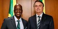 A imagem mostra Sergio Camargo, à esquerda e Jair Bolsonaro, à direita de pé e sorrindo. Eles vestem terno e gravata escuros  Foto: Reprodução/Twitter / Alma Preta