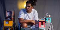 Marcelino é um homem negro que usa dreads. Ele está sentado ao centro da imagem com miniaturas de casas ao redor  Foto: Divulgação/Léu Britto / Alma Preta
