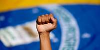 Imagem mostra a bandeira do Brasil ao fundo e a mão fechada de uma pessoa negra para cima.  Foto: Imagem: Reprodução/Sindicato dos Metalúrgicos / Alma Preta
