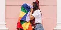 Imagem mostra casal de mulheres se beijando atrás de uma bandeira do arco-íris, símbolo do movimento LGBTQIAP+.  Foto: Imagem: Reprodução/Jéssica Kidermann / Alma Preta