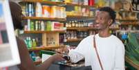 Imagem mostra cliente e caixa de supermercado, ambos homens negros, representando a preferência, apontada por relatório, dos brasileiros por empresas que apoiam a diversidade.  Foto: Imagem: Kampus Production/Pexels / Alma Preta