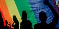 Imagem mostra sombra de pessoas em bandeira LGBTQIAP+.  Foto: Imagem: Pau Barrena/AFP / Alma Preta