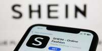 Imagem mostra aplicativo da Shein em celular e logo da marca chinesa ao fundo da imagem  Foto: NurPhoto/Getty Images / BBC News Brasil