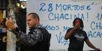 Foto mostra policial e um jovem negro atrás dele, em uma bicicleta, o filmando com um celular.  Foto: André Borges/AFP / Alma Preta