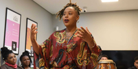 Imagem mostra uma mulher de pele negra, autora do livro ‘Lesbiandade’ centralizada discursando com os braços levemente levantados, em uma sala. A mulher usa um vestido vermelho com detalhes dourados. Ao lado esquerdo, no fundo, mulheres negras sentadas escutando.  Foto: Alma Preta