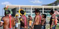 A imagem mostra sete pessoas indígenas lado a lado, sendo somente a segunda delas virada para frente e as outras estão de costas. Eles têm a pele escura, com tom avermelhado, estão sem camisa e vestem adornos tradicionais, como cocar e braceletes. No fundo, a sede do STF, onde acontece o julgamento do Marco Temporal  Foto: Alma Preta