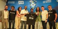 Imagem mostra grupo de pessoas em p&eacute; e sorrindo em frente ao banner da campanha "Com racismo n&atilde;o tem jogo", enquanto seguram uma camisa da iniciativa.  Foto: Alma Preta