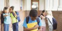 Imagem mostra um menino negro com mochila nas costas e livros nas mãos e outras crianças apontam para ele.  Foto: Alma Preta