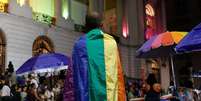 A imagem mostra um homem negro de costa, coberto com a bandeira da população LGBTQIA+  Foto: Alma Preta