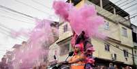 Imagem mostra a performance de Milu Almeida e Luiz Otávio na favela da Nova Holanda (RJ). Na foto, os artistas aparecem em cima de uma moto com trajes na cor rosa e laranja, enquanto dezenas de pessoas os cercam.  Foto: Alma Preta