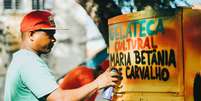 A imagem mostra um homem negro com uma lata de tinta em frente a uma geladeira com os dizeres &ldquo;Gelateca Cultural Maria Bet&acirc;nia de Carvalho&rdquo;.  Foto: Alma Preta