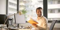 Homem sorri no escritório com livro nas mãos  Foto: Pexels | Banco gratuito de imagens