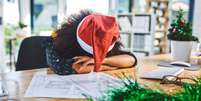 mulher com chapeu de papai noel descansa a cabeça em mesa de escritório  Foto: Getty Images / BBC News Brasil