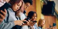 Quatro alunos de pé contra uma parede de tijolos, sem falar uns com os outros enquanto cada um olha para um telefone em suas mãos  Foto: Getty Images / BBC News Brasil