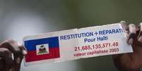 Manifestante segura um cartaz em francês com os dizeres “Restituição e compensação para o Haiti” enquanto ouve o discurso do presidente francês François Hollande do lado de fora do Palácio Nacional em Porto Príncipe, Haiti, em maio de 2015. (Imagem ilustrativa)  Foto: AP - Dieu Nalio Chery / RFI