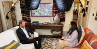 O príncipe Harry está sentado em um sofá branco na sala de estar, em frente a Nada Tawfik que está sentada em um sofá laranja listrado. O equipamento de iluminação da TV pode ser visto acima de suas cabeças.  Foto: BBC News Brasil