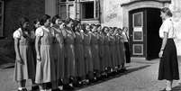 Meninas uniformizadas enfileiradas em frente a uma mulher em foto em preto e branco Foto: Alamy / BBC News Brasil