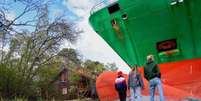 Três pessoas, vestindo jeans e jaquetas casuais, estão de pé, observando um enorme navio porta-contêineres verde que encalhou em terra firme. Uma casa parece pequena e está ao lado do navio. Foto: EPA / BBC News Brasil
