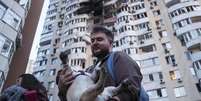Um homem carrega seu cachorro em frente a um prédio residencial de vários andares danificado após um ataque de drone russo em Kiev, Ucrânia, nesta sexta-feira  Foto: AP - Evgeniy Maloletka / RFI