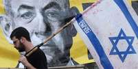Um homem com uma bandeira israelense passa por um cartaz com a foto de Netanyahu  Foto: JACK GUEZ/AFP via Getty Images / BBC News Brasil