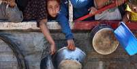 Uma menina palestina encostada em um muro, ao lado de outras pessoas, segurando uma tigela de metal, à espera da distribuição de sopa de lentilha na Cidade de Gaza, no norte da Faixa de Gaza  Foto: AFP / BBC News Brasil