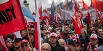 Manifestantes seguram bandeiras nacionais da Polônia durante um protesto antimigração convocado pelo partido nacionalista Lei e Justiça, na Praça do Castelo Real, em Varsóvia, em 11 de outubro de 2025.  Foto: AFP - WOJTEK RADWANSKI / RFI